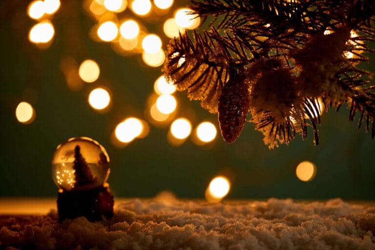 Moody Christmas Decor with warm golden lights, evergreen branches, pinecones, and a small snow globe on a cozy winter surface